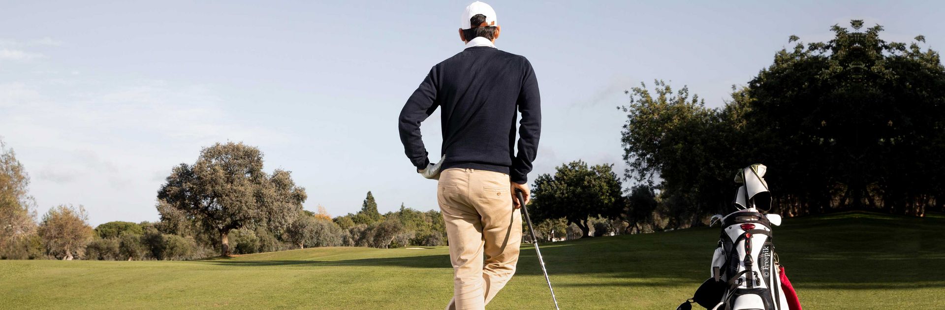 Golfer steht entspannt mit dem Rücken zum Fotografen und Blickt in die Ferne. Neben ihm steht das Golfbag.