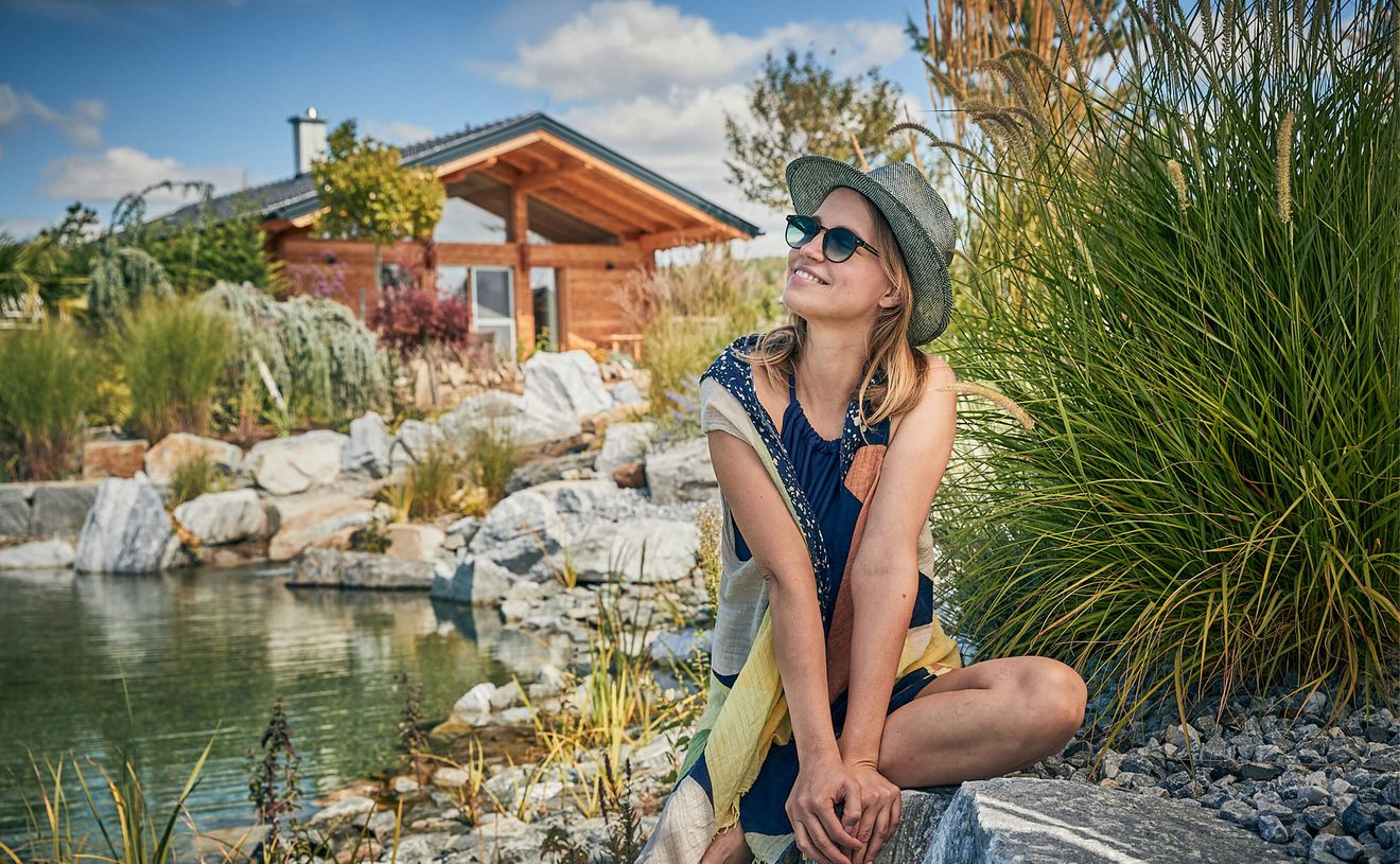 Eine Frau mit Sonnenbrille und Hut sitzt auf einem Stein am Ufer des Gartensees. Im Hintergrund ist das Chalet Typ Landhaus zu sehen.