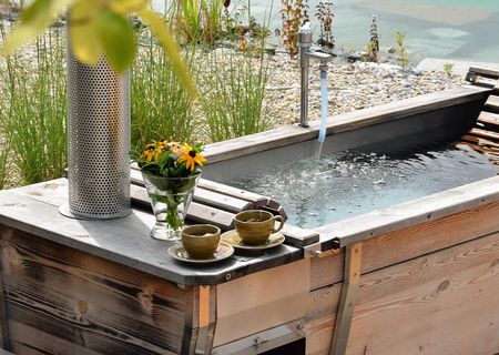 Eine dampfende Feuerwasserwanne aus Holz mit zwei Teetassen und einem Blumenstrauß lädt zum Entspannen ein. Direkt am Ufer gelegen, verschmilzt das naturnahe Wellness-Erlebnis mit der ruhigen Gartenlandschaft.