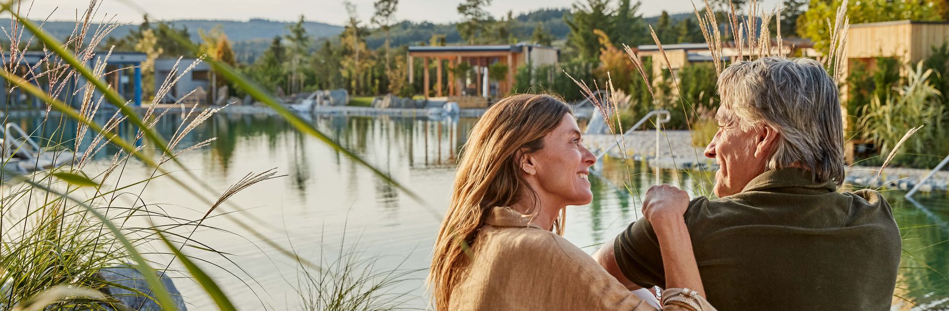 Ein Paar sitzt am Ufer des Gartensees. Im Hintergrund sind die verschiedensten Chalets zu sehen.